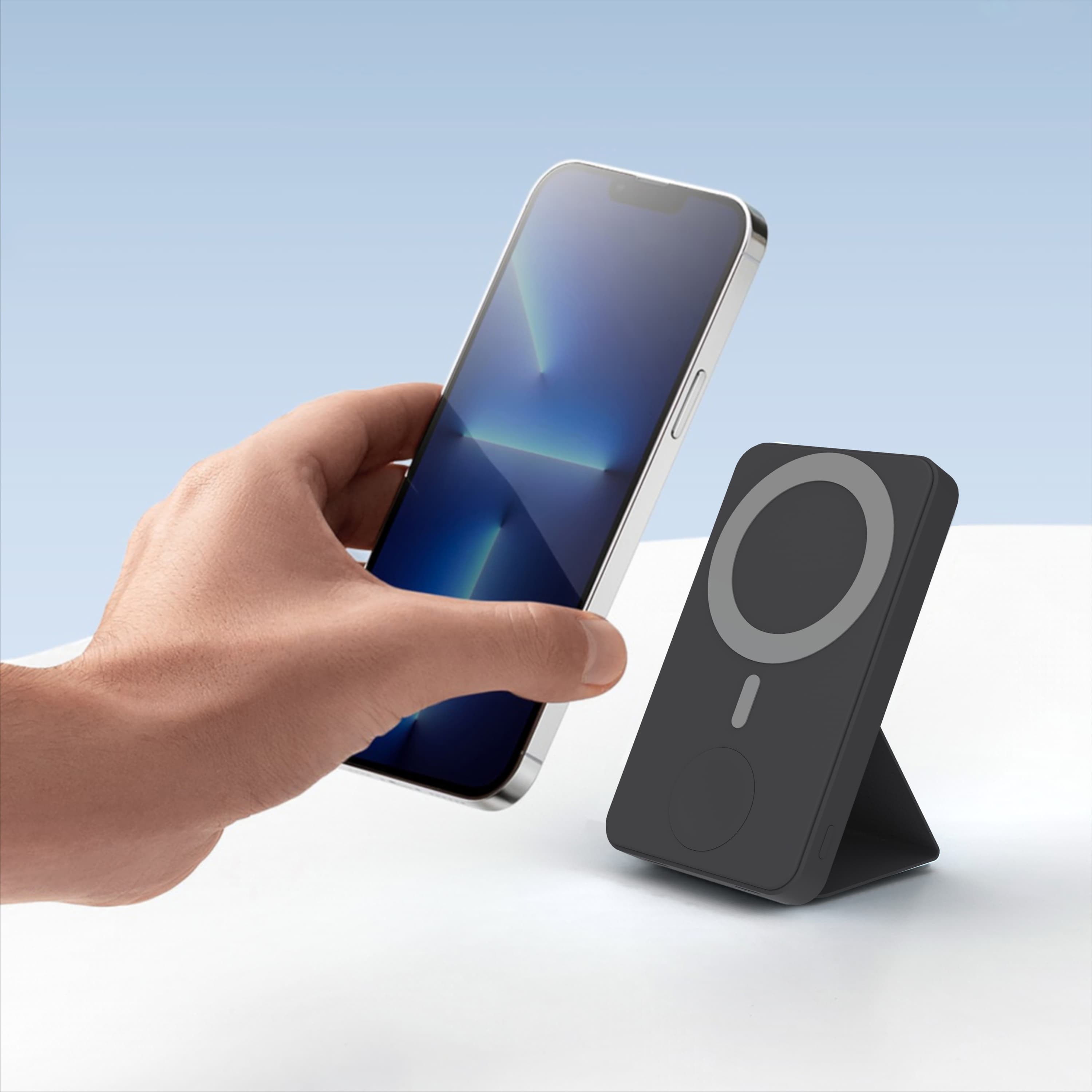 Hand holding a smartphone near a black magnetic wireless power bank with an integrated stand.