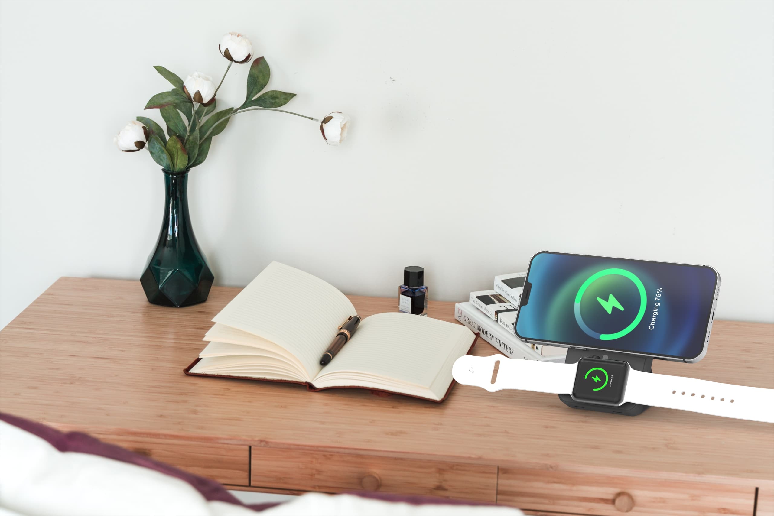 Smartphone and smartwatch charging on a wooden desk next to an open notebook and vase.